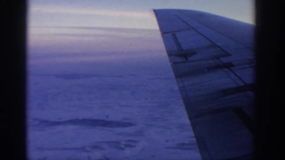 1963 home-movie frame with an airplane wing at right, a dark window border, and clouded terrain under cool blue light.