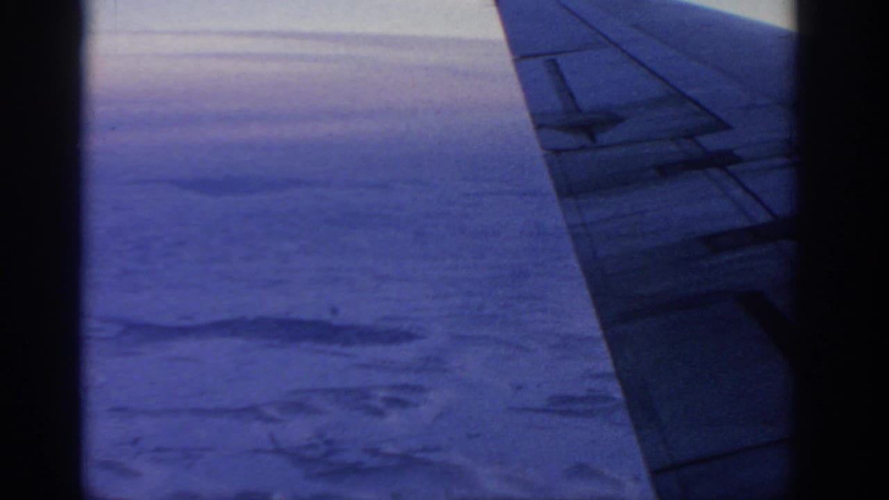 1963 8mm view from an airplane window near Warsaw, with the wing crossing a blue-toned sky above clouds and land.