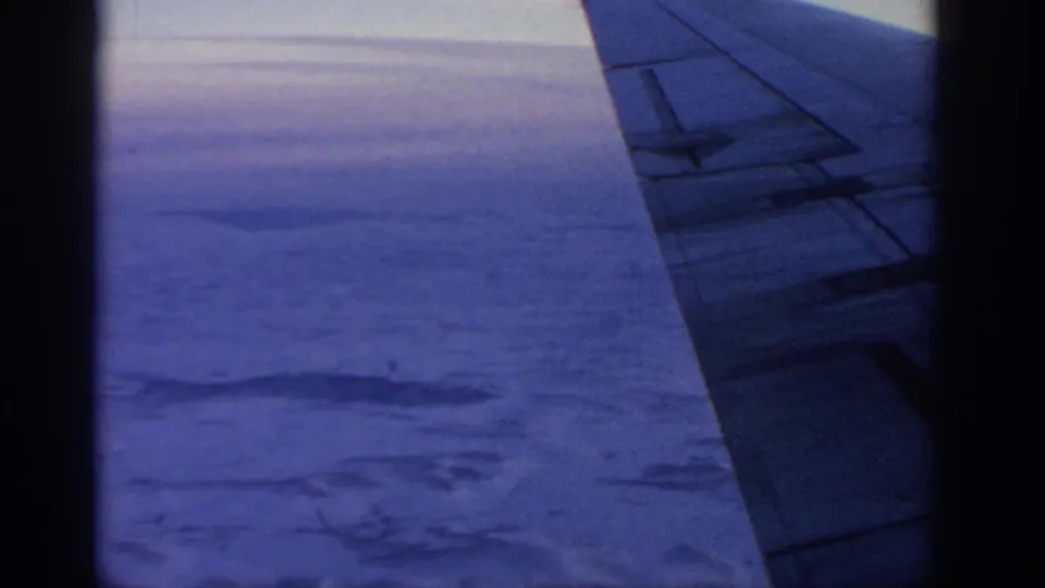 1963 8mm view from an airplane window near Warsaw, with the wing crossing a blue-toned sky above clouds and land.