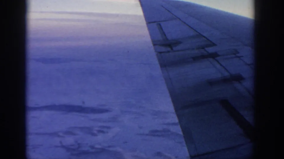 Blue-toned 8mm frame of an airplane wing over cloud layers and land below, filmed from a passenger window.