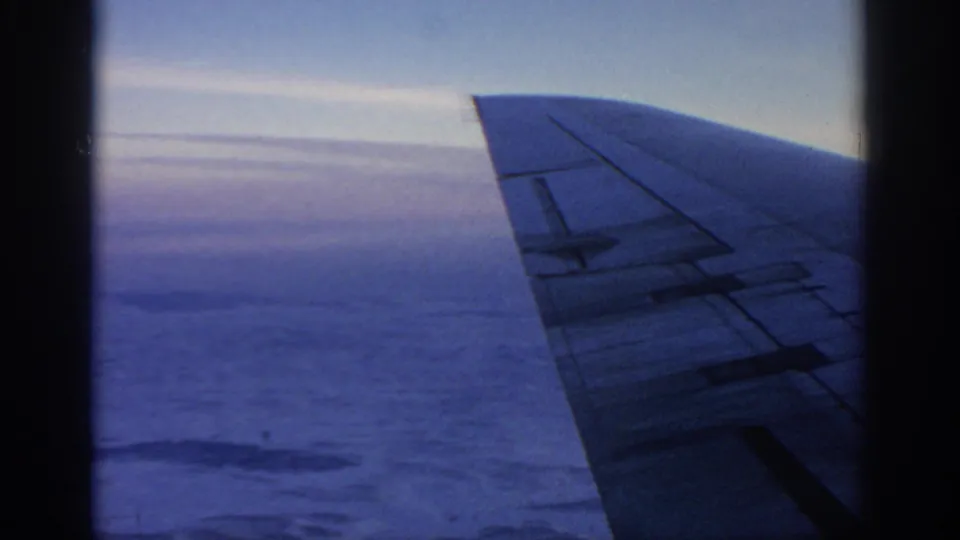 8mm passenger view of an aircraft wing against a cool sky, with cloud cover and indistinct ground visible below.