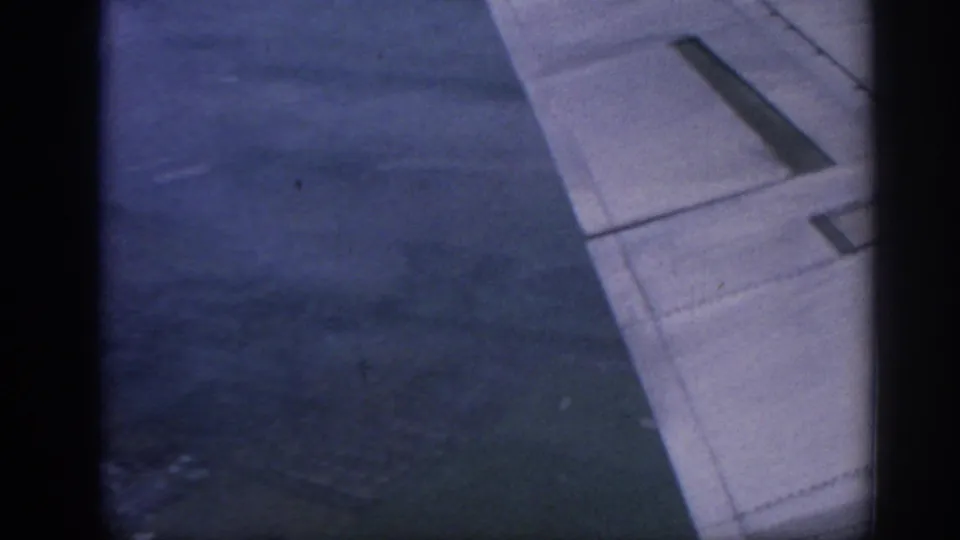 Closer 8mm view of the airplane wing and window edge, with blurred earth tones visible through the haze below.