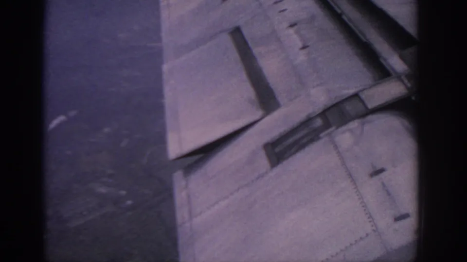 Late-frame 8mm view of the aircraft wing and flap detail, partly bordered by the dark airplane window frame.