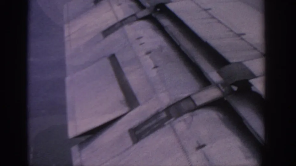 1963 in-flight 8mm view showing the aircraft wing and mechanical flap area against a hazy purple sky.
