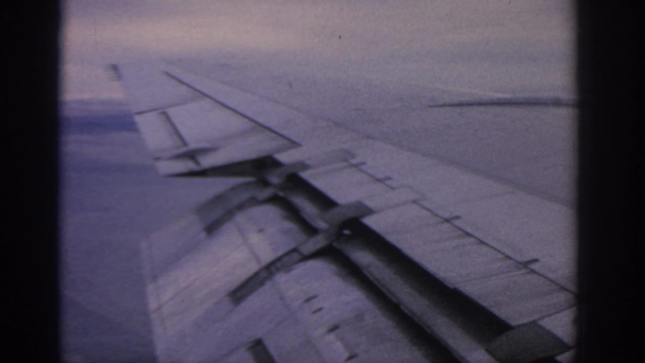 1963 8mm passenger-window view near Warsaw metadata, showing an airplane wing framed by dark window edges and a soft blue-purple sky.