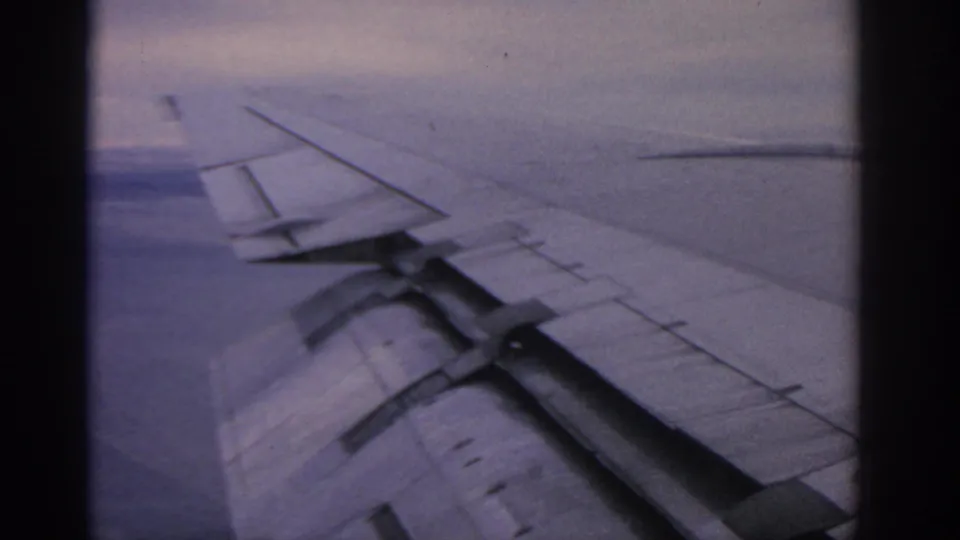 1963 8mm passenger-window view near Warsaw metadata, showing an airplane wing framed by dark window edges and a soft blue-purple sky.