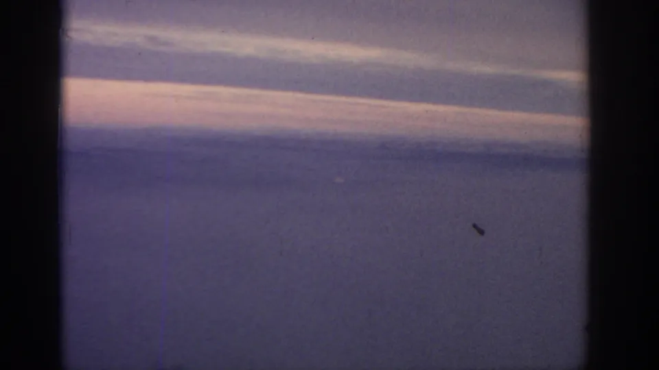 In-flight 8mm view over cloud layers and distant horizon, with the aircraft window frame visible and no landmarks identifiable.