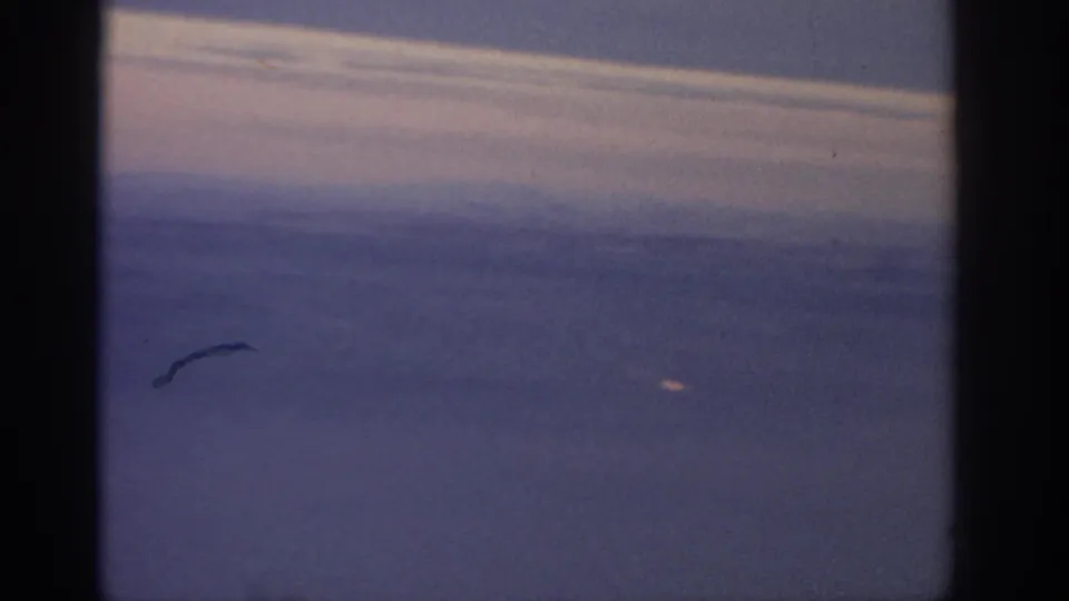 Vintage 8mm in-flight view of cloud bands and pale sky, framed by the airplane window with visible scratches and dust.