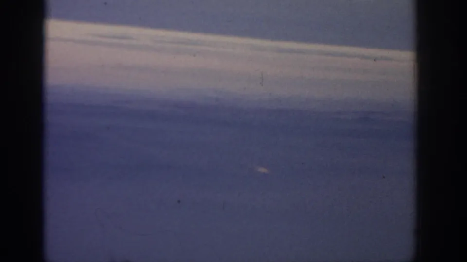 Soft 8mm aerial view over blue water with a cloud band near the horizon, dark film borders, and minor dust marks.