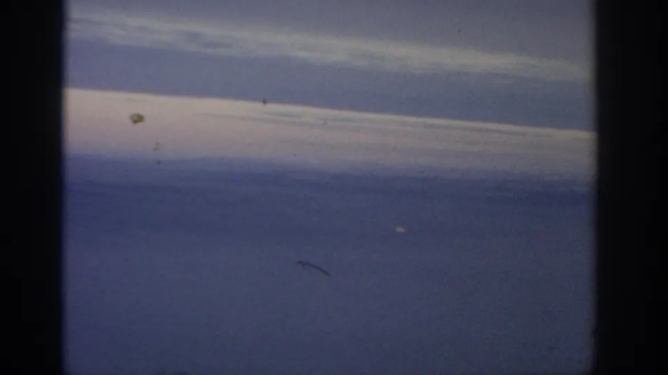 1960s 8mm-style frame showing slanted pale cloud layers over blue water, with grain, dust, scratches, and dark frame edges.