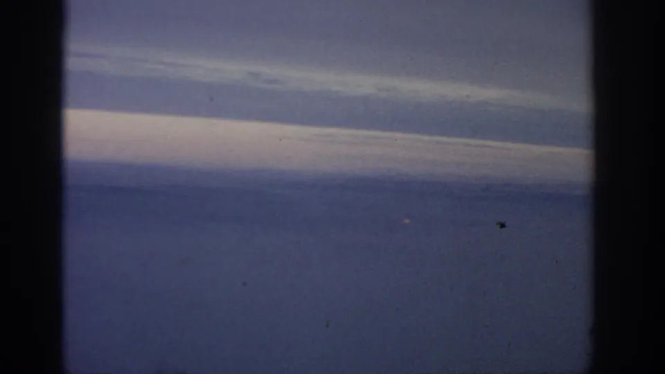 Aerial-looking 8mm view of blue water beneath a pale cloud band, with dark film borders, grain, and small dust marks.