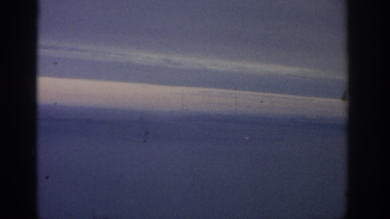 1963 8mm passenger view near Warsaw, showing pale cloud bands over a hazy blue landscape through an airplane window.