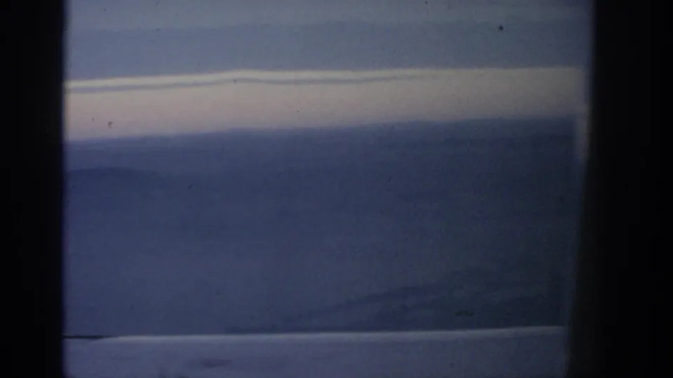 A 1963 airplane-window frame over the Warsaw area, with soft blue haze, a pale cloud streak, and dark cabin-window edges.