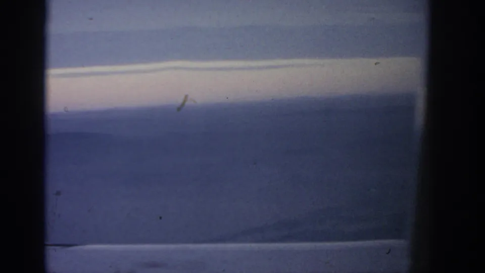 1963 8mm travel footage frame with a pale cloud band, blue-violet haze, and low landscape seen from an airplane window.