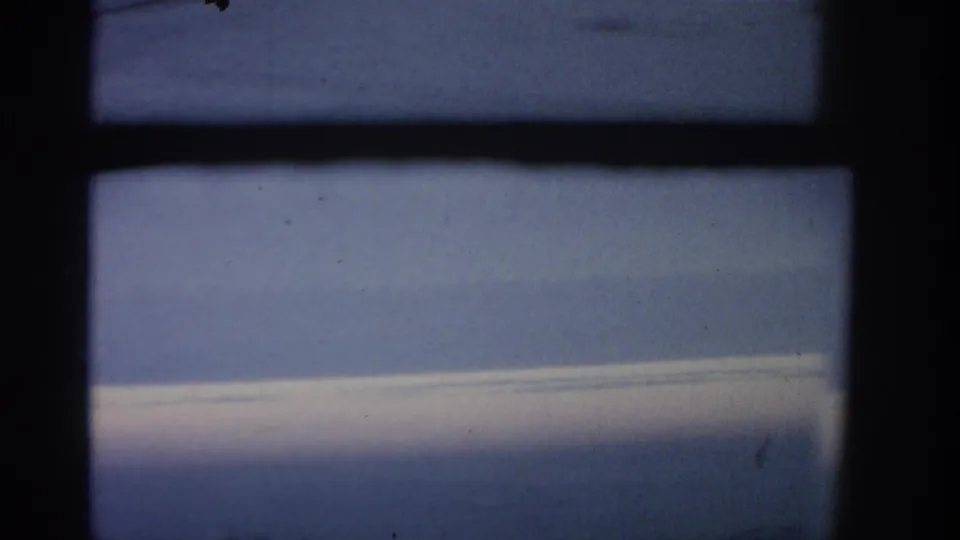 1963 8mm flight view associated with Warsaw metadata, seen through a dark airplane window frame toward blue sky and clouds.