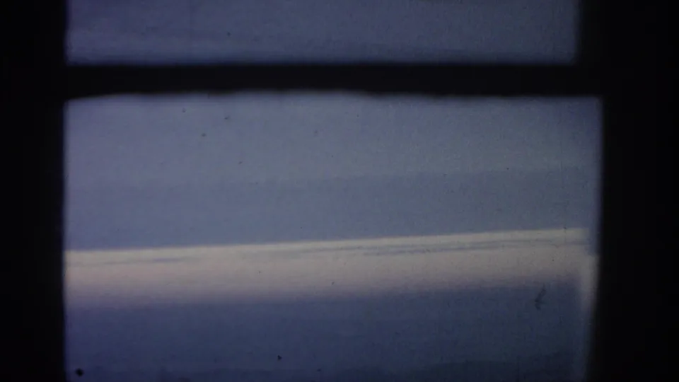 A 1963 home-movie frame shows the lower airplane window area with blue sky, a cloud shelf, and dark framing.
