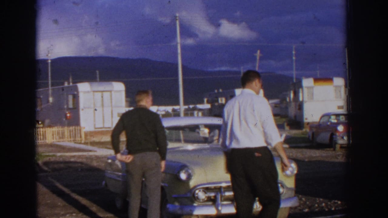 Two men walk near a pale classic Chevrolet in a trailer-park setting with mountains behind.