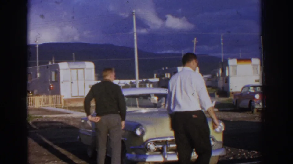Zurich, Switzerland home-movie frame from 1963 showing two adult men beside a pale classic Chevrolet near mobile homes and distant mountains.