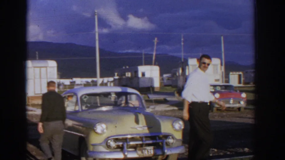 Zurich, Switzerland 1963 frame with a man in sunglasses smiling toward the camera beside a pale classic Chevrolet at a trailer park.