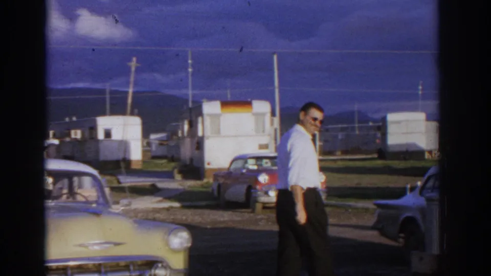 1963 Zurich home-movie frame showing a man in sunglasses walking near the front of a classic Chevrolet with trailer-park structures behind.