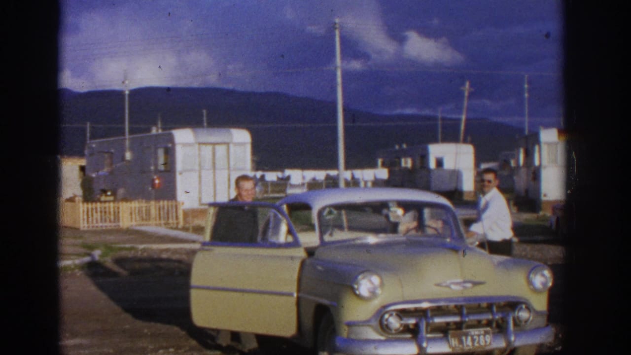 1963 home-movie frame from Zurich, Switzerland showing two men near a yellow and white classic car among trailers and distant mountains.