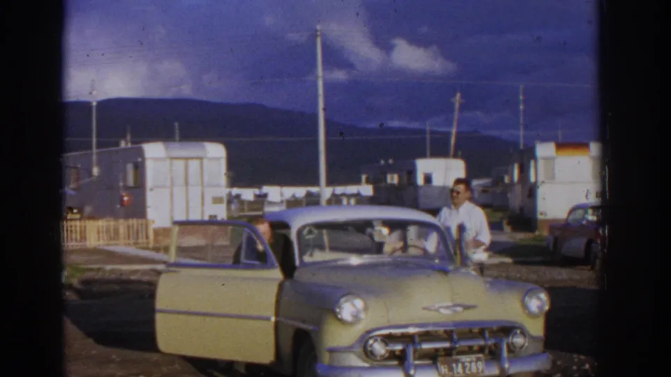 1963 Zurich campground frame with a man by an open yellow car door, seated occupants, trailers, poles, and mountains in the background.