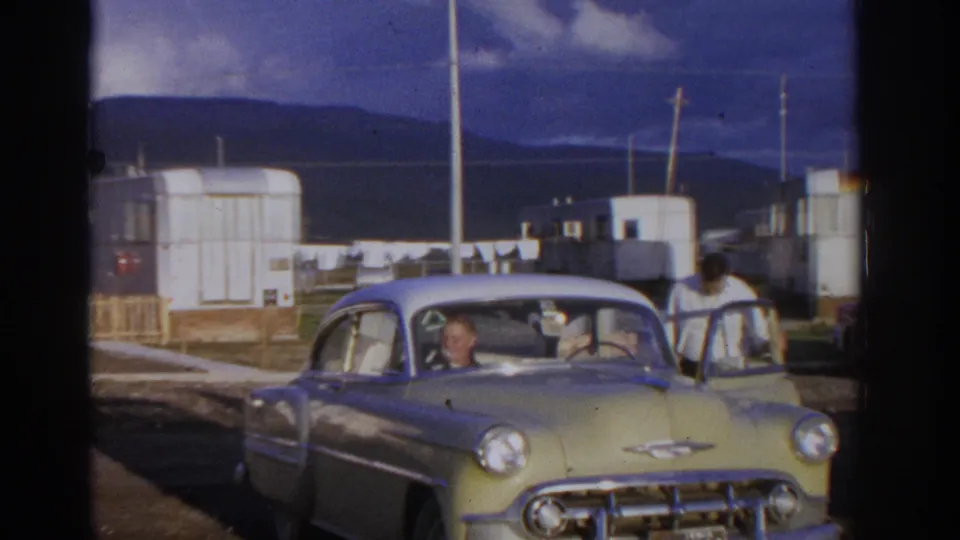 Home-movie frame from 1963 Zurich showing a yellow and white classic car at a campground with men inside and trailers behind it.