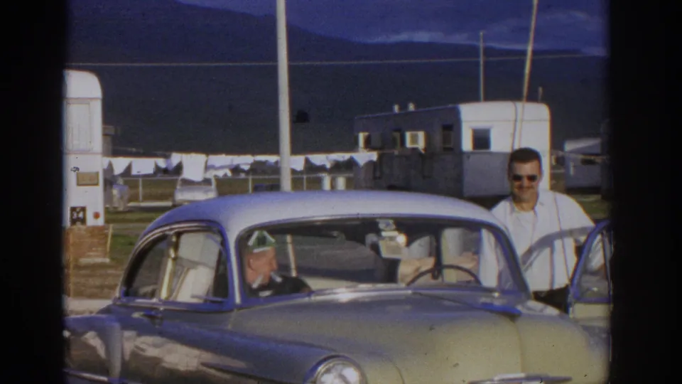 1963 Zurich frame showing a man in sunglasses beside a yellow and white classic car, with seated occupants, trailers, laundry, and mountains behind.