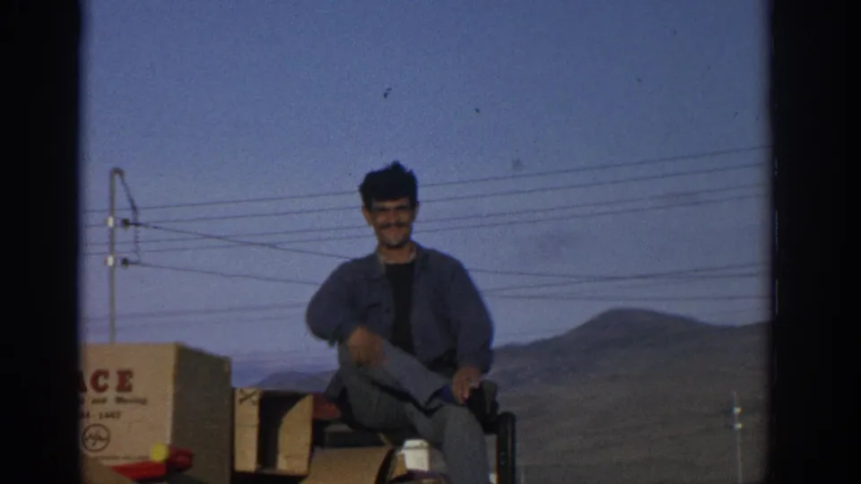 1963 home-movie frame of a seated man on stacked moving boxes, with rural hills, wires, and visible 8mm frame edges.