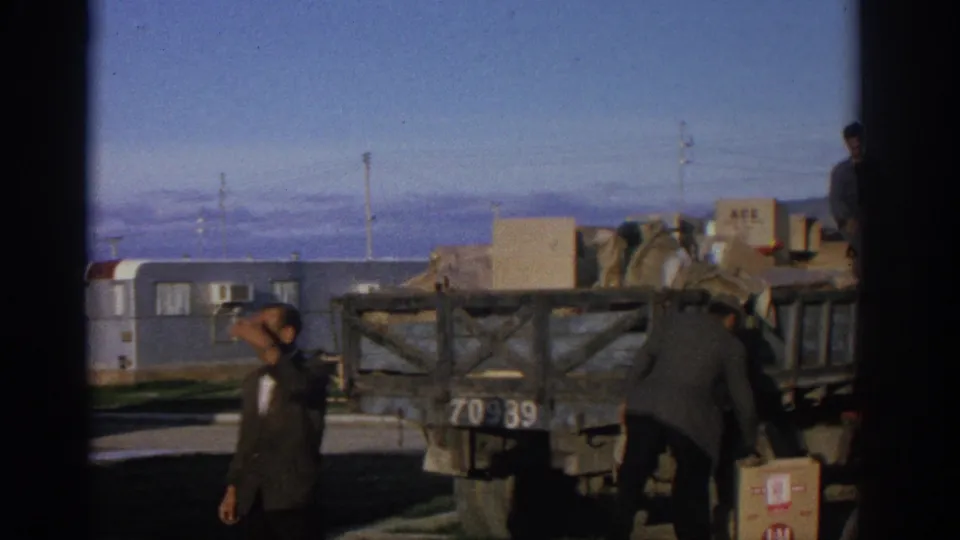 Wide 8mm frame from Zurich-area footage showing men beside the rear of a loaded truck, boxes, a trailer, and distant mountains.