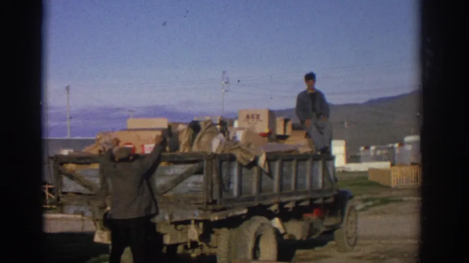 1963 Zurich home-movie still of a man reaching up at the back of a wooden truck stacked with cardboard boxes and bags.
