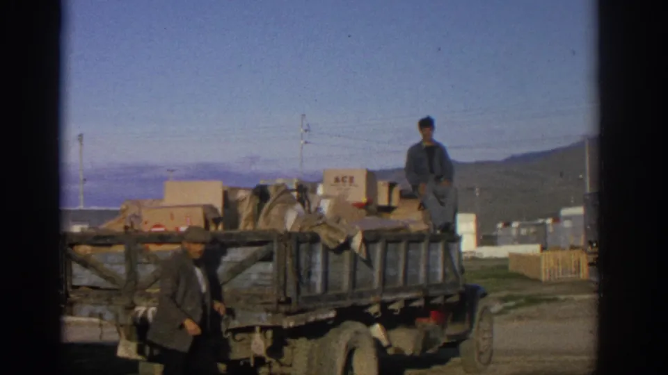 1963 Zurich frame showing a loaded flatbed truck, one man walking in front, and another man seated among boxes and bundles.