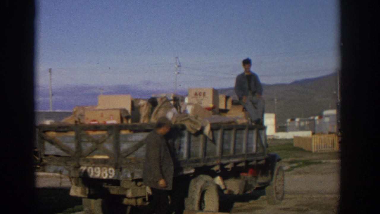 1963 Zurich home-movie frame showing one man seated atop a loaded wooden truck while another stands near the truck bed.