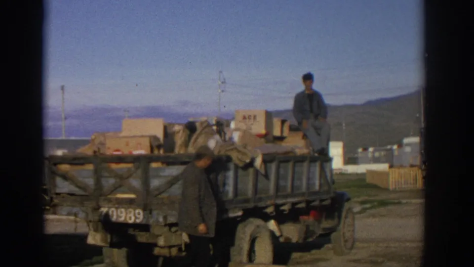 1963 Zurich home-movie frame showing one man seated atop a loaded wooden truck while another stands near the truck bed.