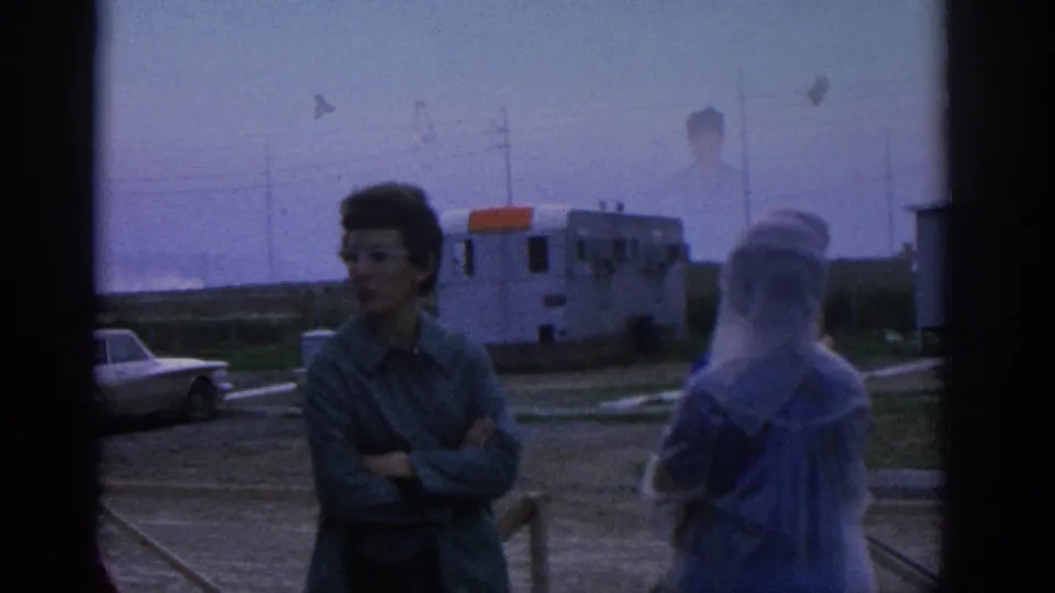 1963 Zurich home-movie frame showing adults in conversation beside an open area, with a parked car and low building behind them.