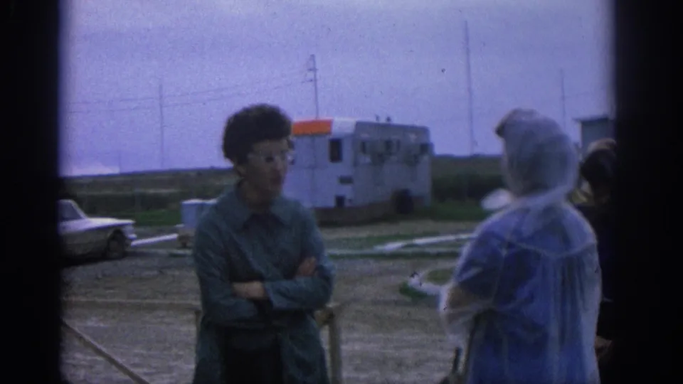 1963 Zurich frame showing a man with arms crossed speaking with older adults in an open roadside setting.