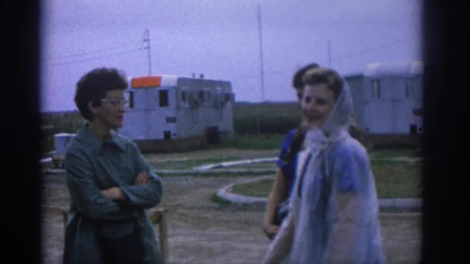 1963 Zurich frame with women in head coverings and a woman in glasses sharing a casual outdoor exchange near trailers.