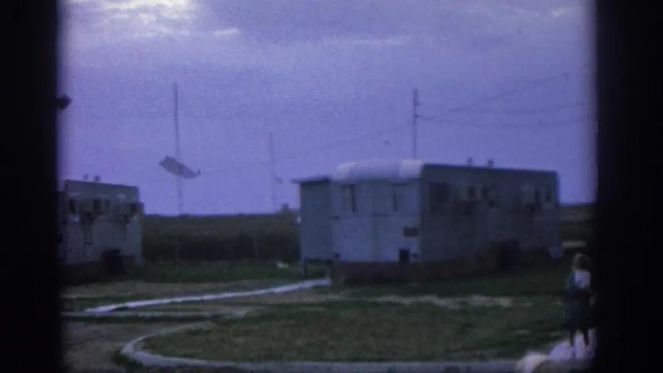 1963 Zurich frame showing mobile trailers, utility poles, open grass, and two people partly visible at the right edge.