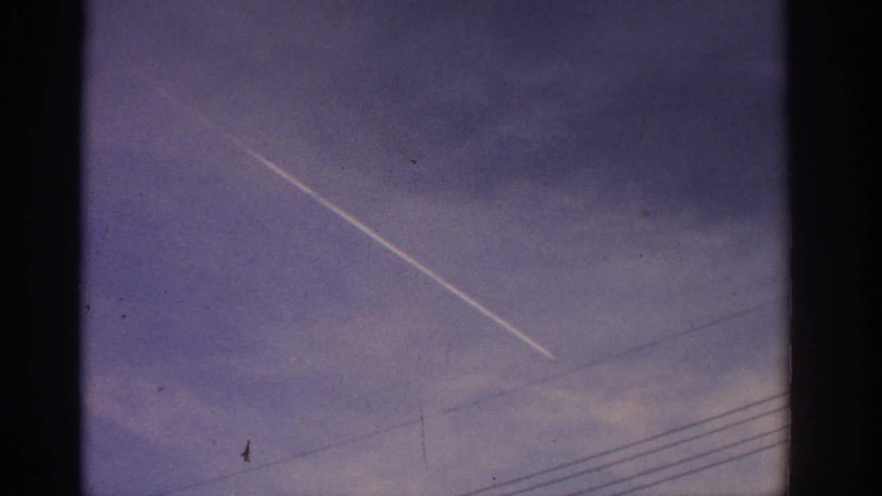 Blue sky with a diagonal white contrail, faint cloud bands, wires, and dark film borders.