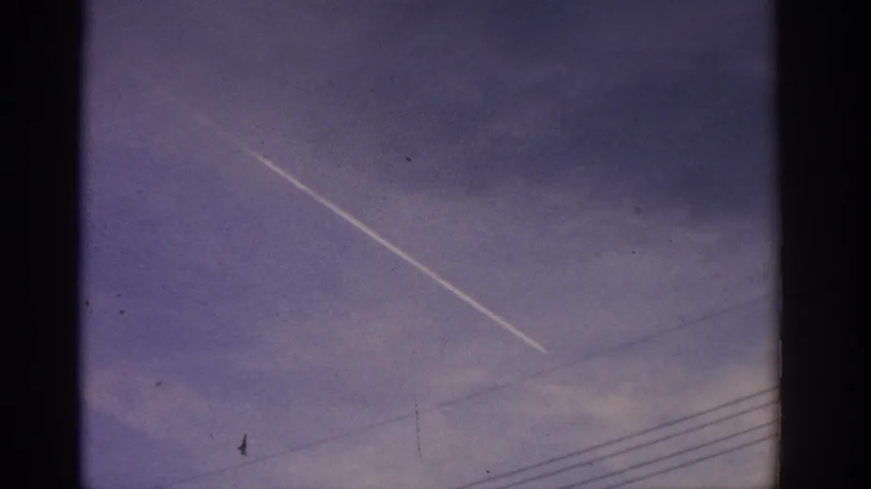 1963 Zurich home-movie frame showing a white contrail crossing blue-purple sky above faint overhead wires.
