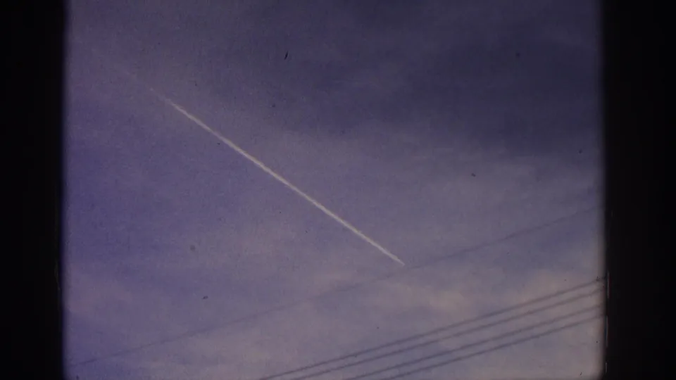 Home-movie view of a diagonal airplane contrail over Zurich sky, framed by dark 8mm gate edges and utility wires.