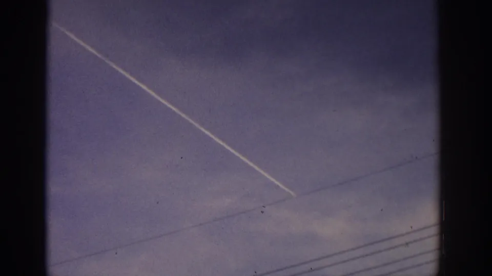 1960s 8mm frame of a bright contrail above faint wires, with soft color and visible film texture.
