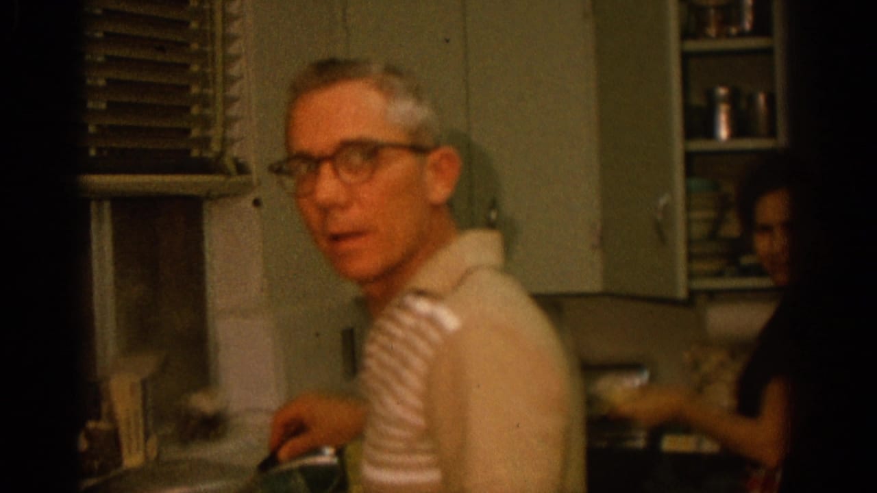 Man in glasses turns from a stove in a small kitchen while a woman works behind him.