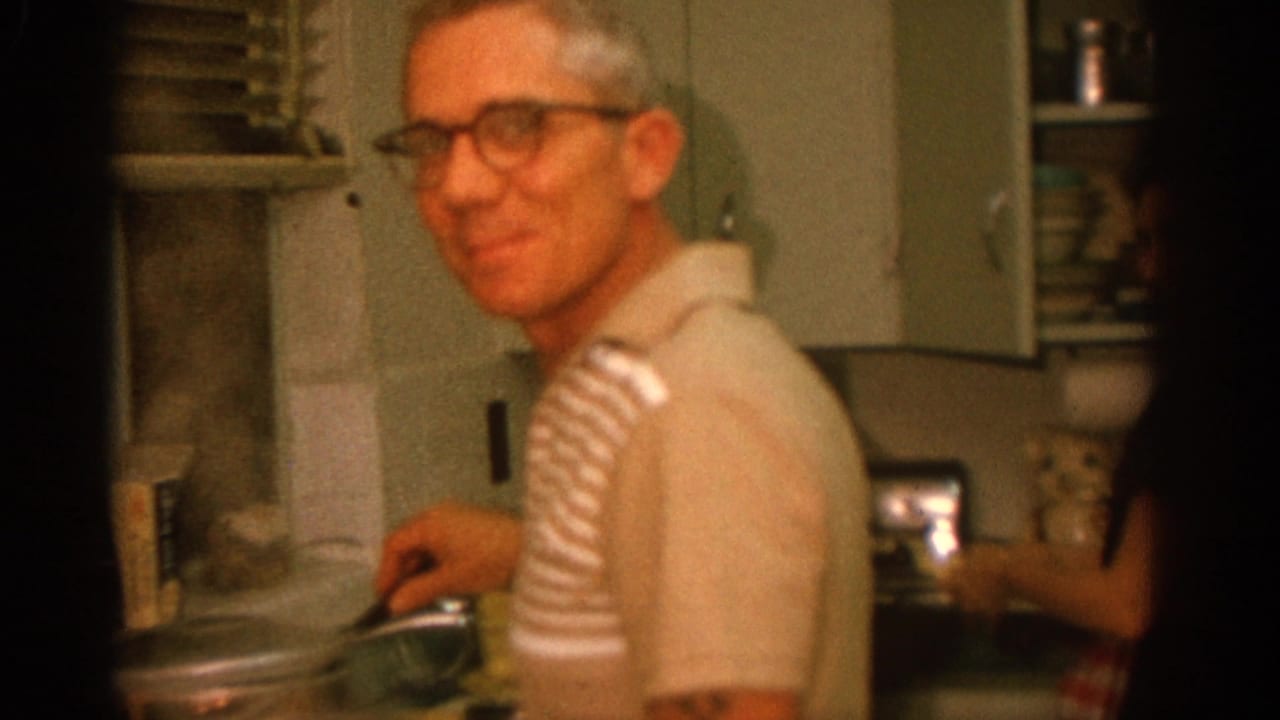 Smiling man at the stove with a woman partly visible at the kitchen counter.