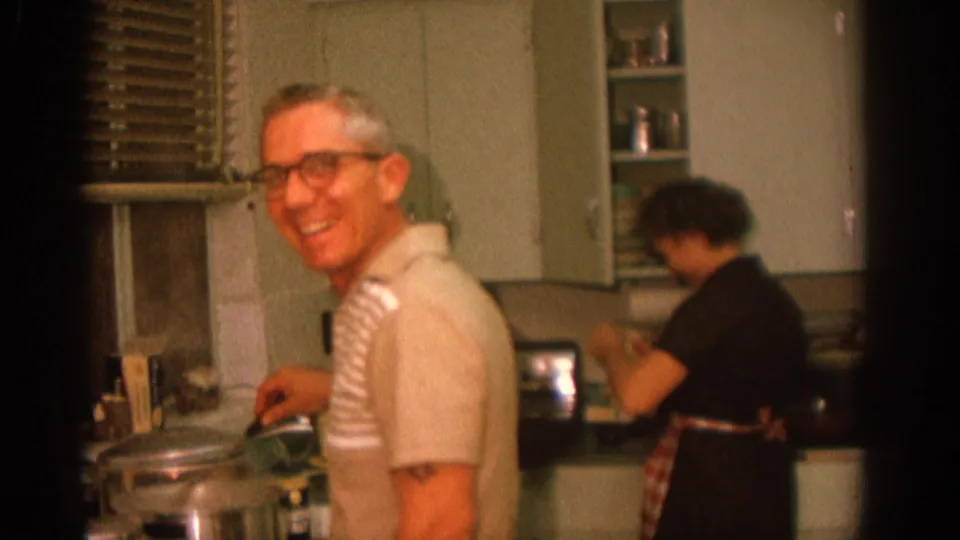 The man laughs or smiles broadly beside the stove in the Zurich kitchen as the woman continues preparing food at the counter.