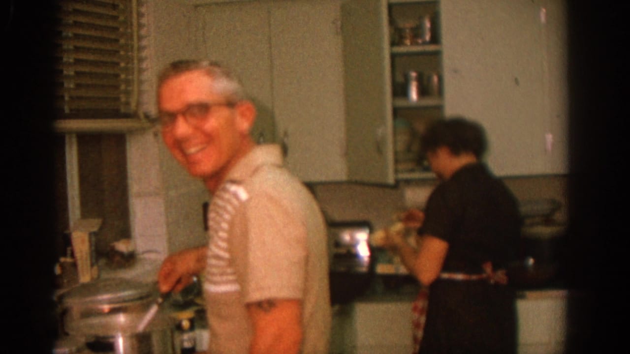 Man in glasses smiles while cooking at a stove as a woman works nearby in a kitchen.