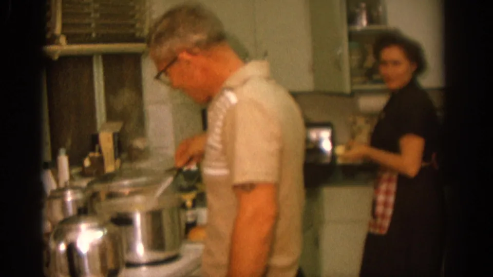 A man tends cookware at a stove in a 1963 Zurich kitchen while a woman in a dark dress and checked apron stands nearby.