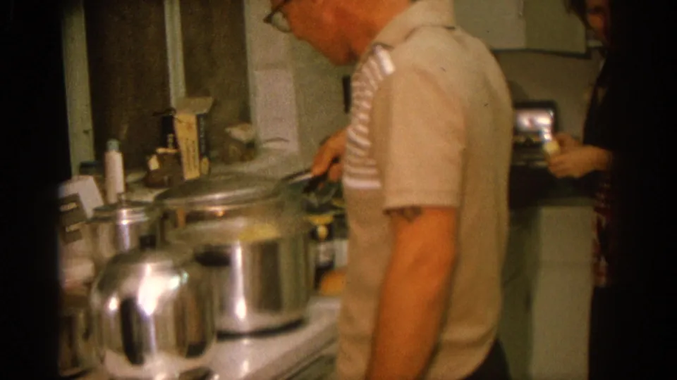 Close 8mm frame of a man cooking in a Zurich kitchen, with large metal pots and a kettle-like vessel filling the foreground.
