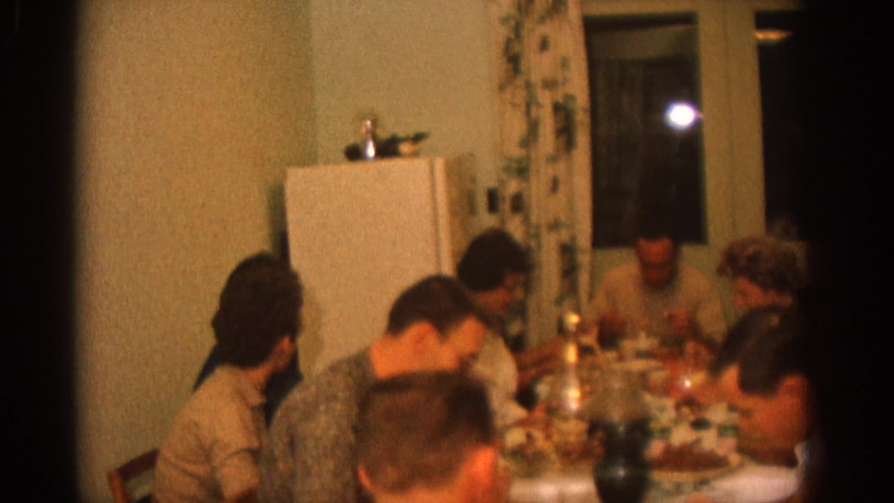 Adults seated around a dining table in a warm interior with a refrigerator and curtains behind them.