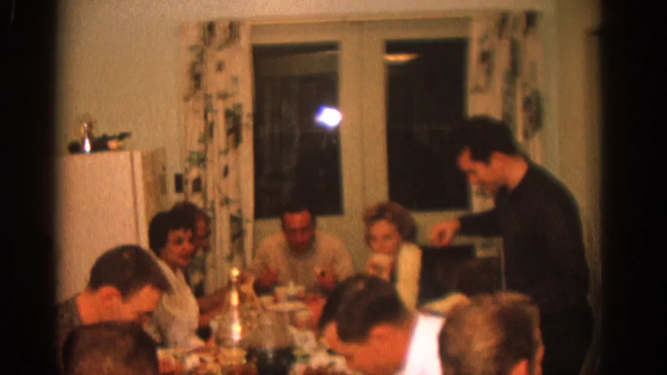 1963 Zurich home-movie still of a man serving at a crowded family dinner table in a warm private interior.